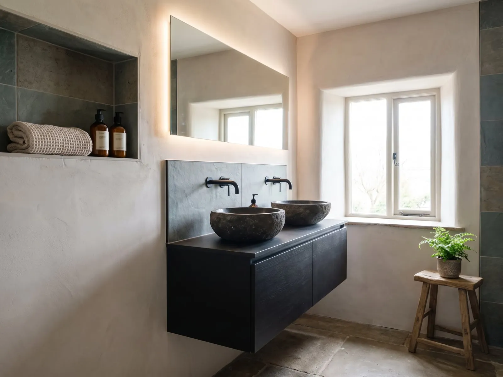 Vanity unit in black with two round dark stone basins, black wall-mounted taps and a wide lit mirror with recesses beside it