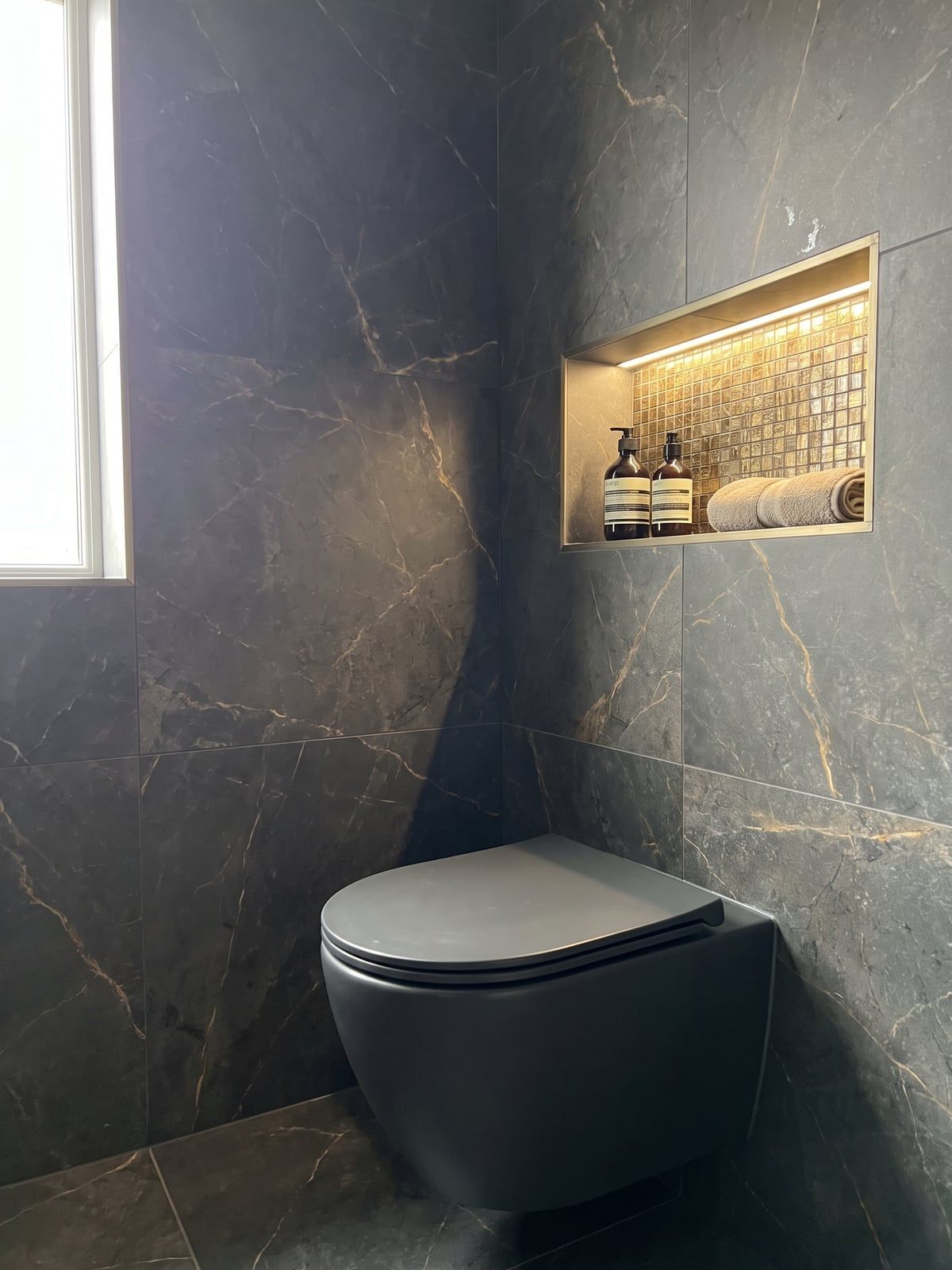 Matt anthracite wall-hung WC against dark marble-effect tiles with gold veining, with a lit recess in gold-toned tile beside it
