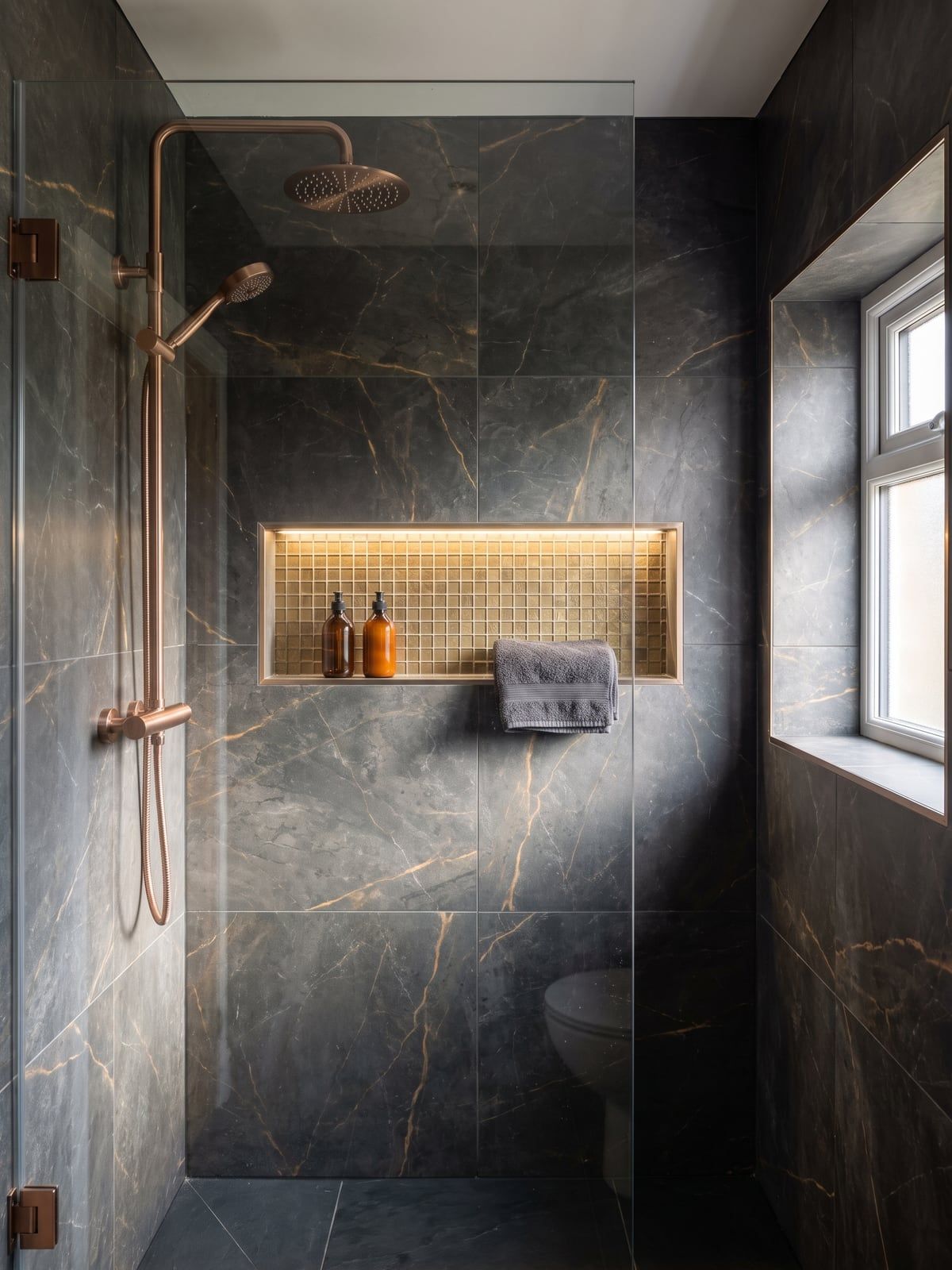 Shower area in dark marble effect with a brushed copper shower set and a lit recess in gold-toned tile