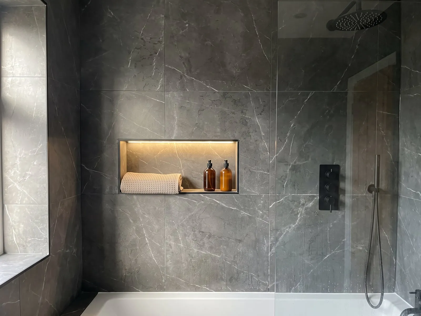 Shower wall in large dark marble-effect tiles with a lit recess, black concealed valves and the edge of a bath
