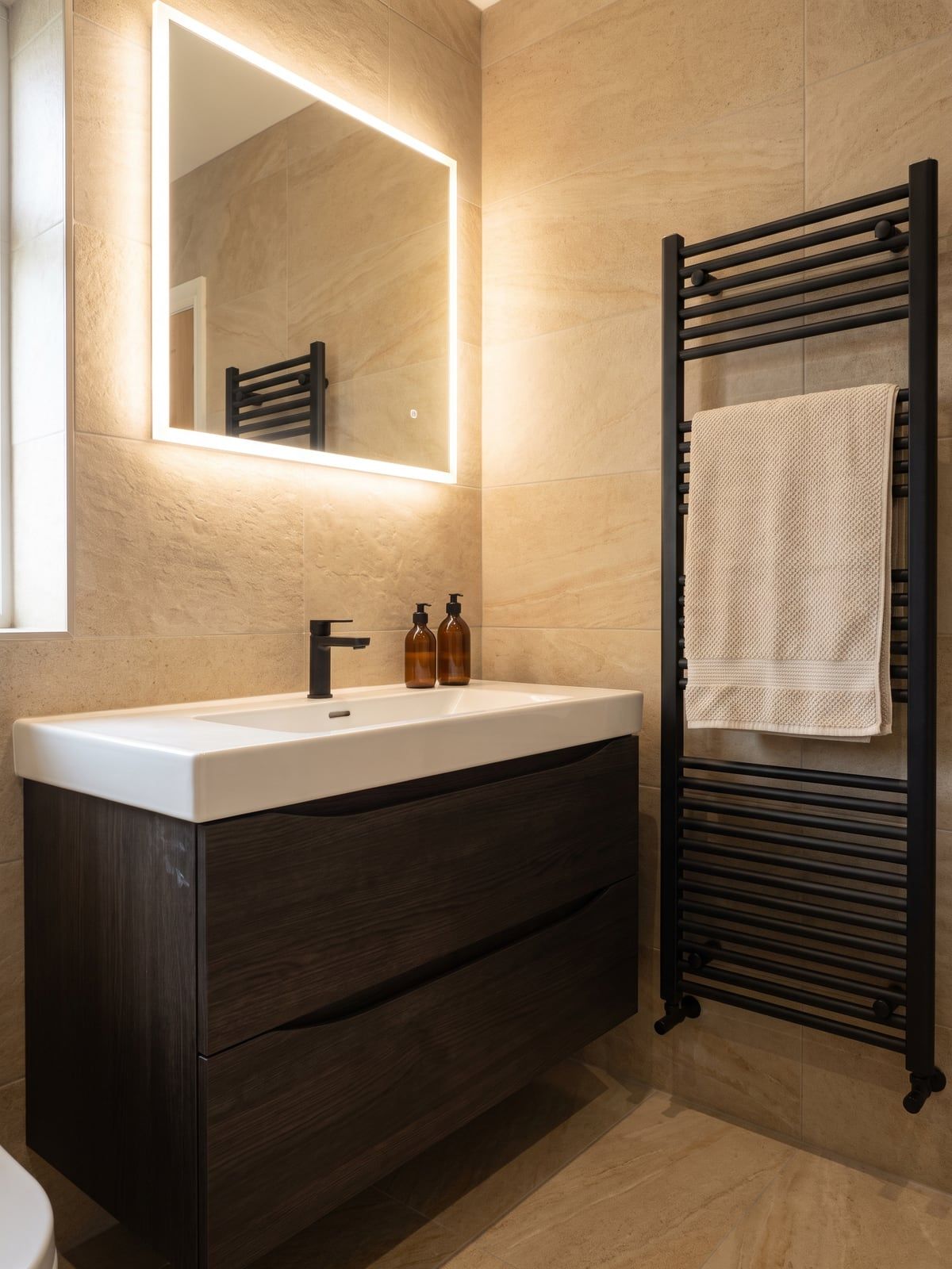 Dark vanity unit with a wide basin and a backlit mirror above it, beside a matt black towel radiator