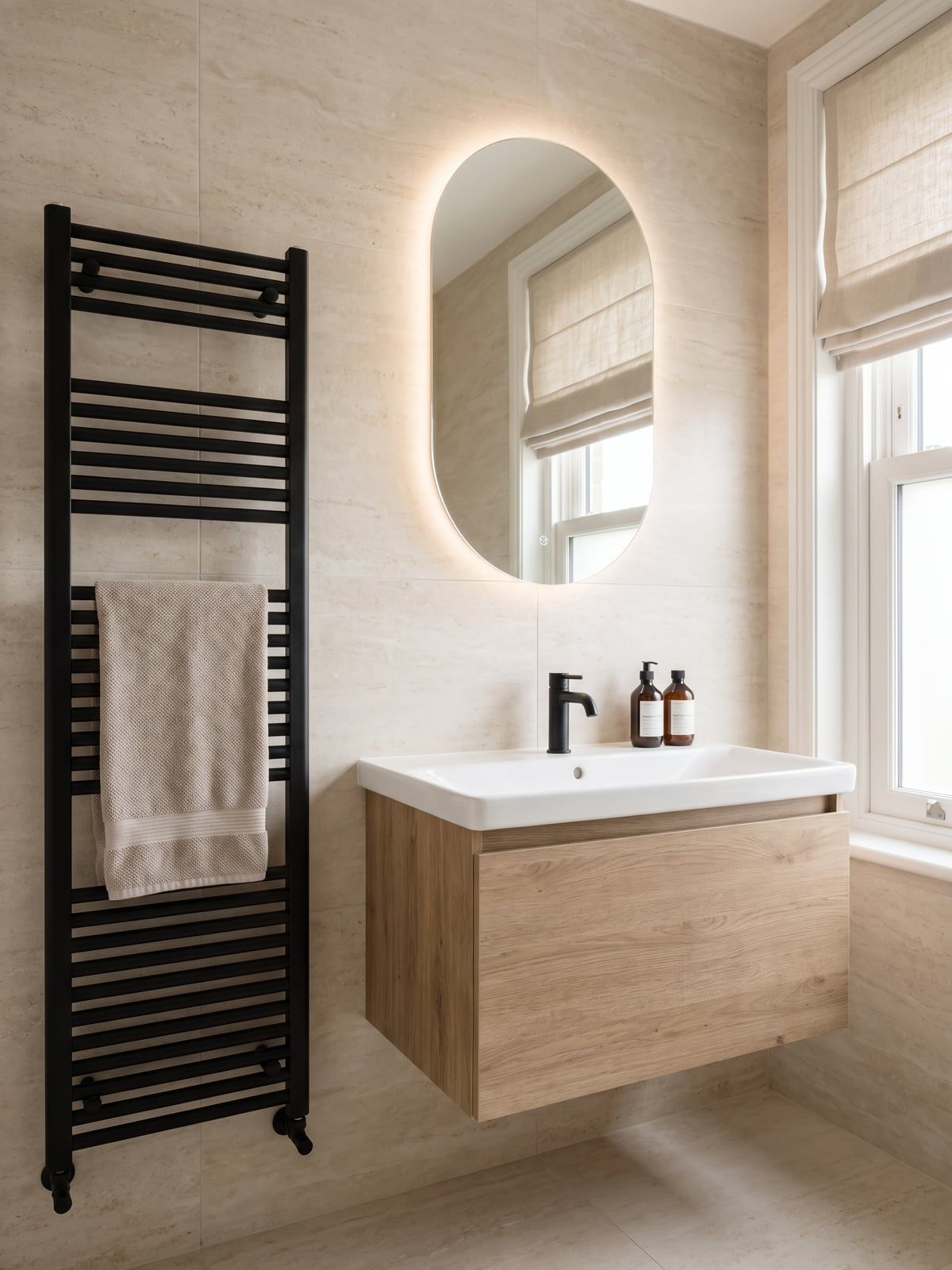 Bathroom with large travertine-effect tiles, a tall matt black towel radiator, an oval backlit mirror and an oak-effect vanity unit with a wide white basin