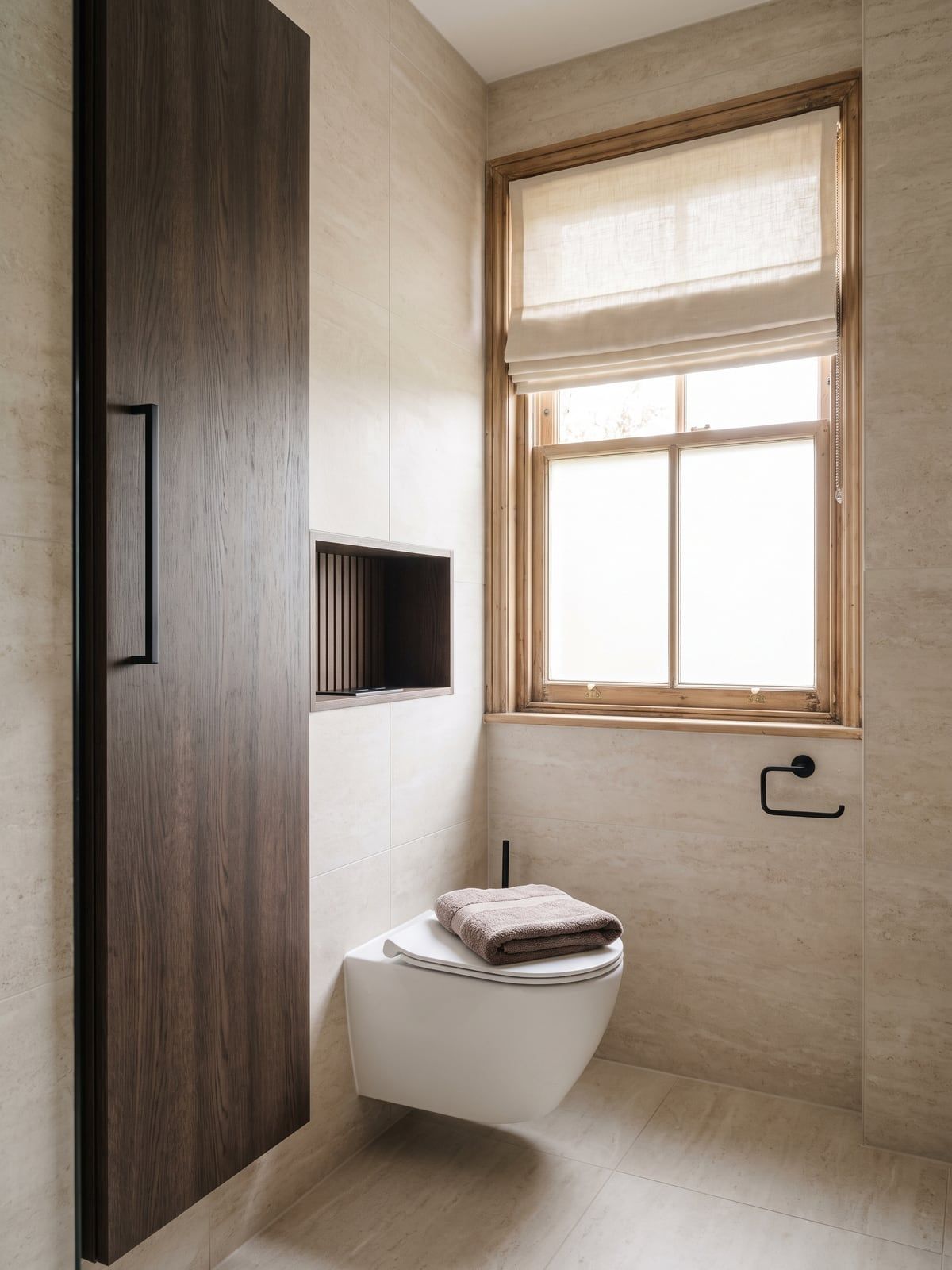 Corner of the bathroom with a tall dark timber-effect cabinet, the sash window with a roman blind and the wall-hung WC beside it
