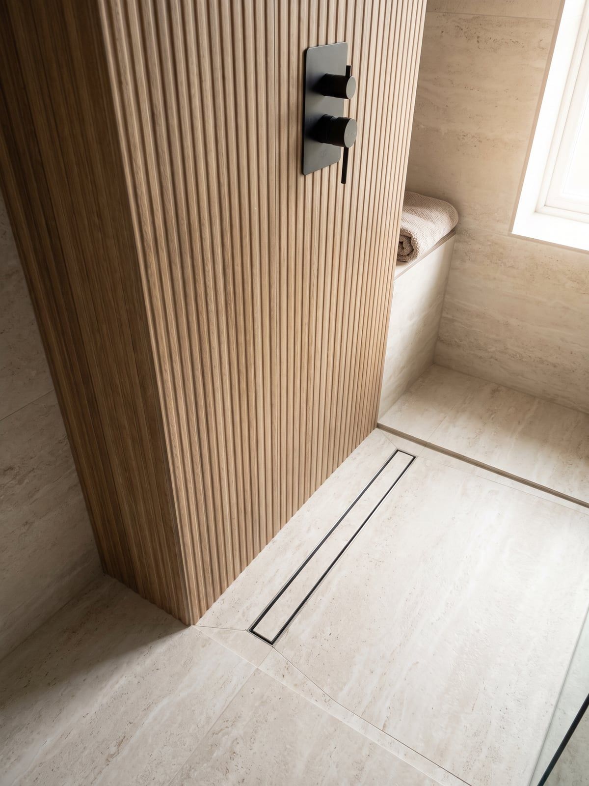 Shower floor with a long dark linear drain, travertine-effect tiles running through and a matt black thermostatic valve on the fluted oak-effect wall