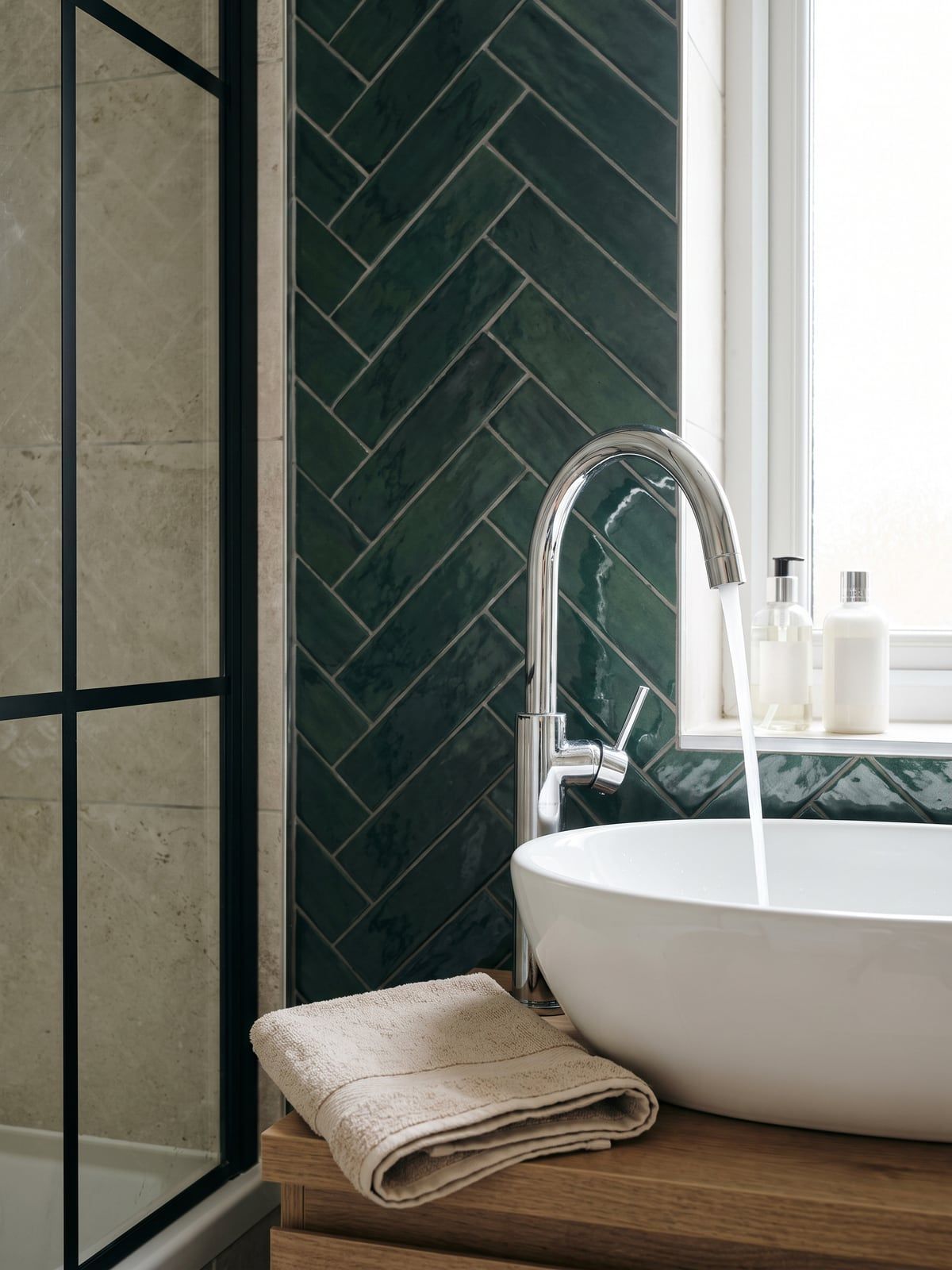 Basin with a tall curved chrome tap running, with the green herringbone wall and the black-framed shower screen behind it