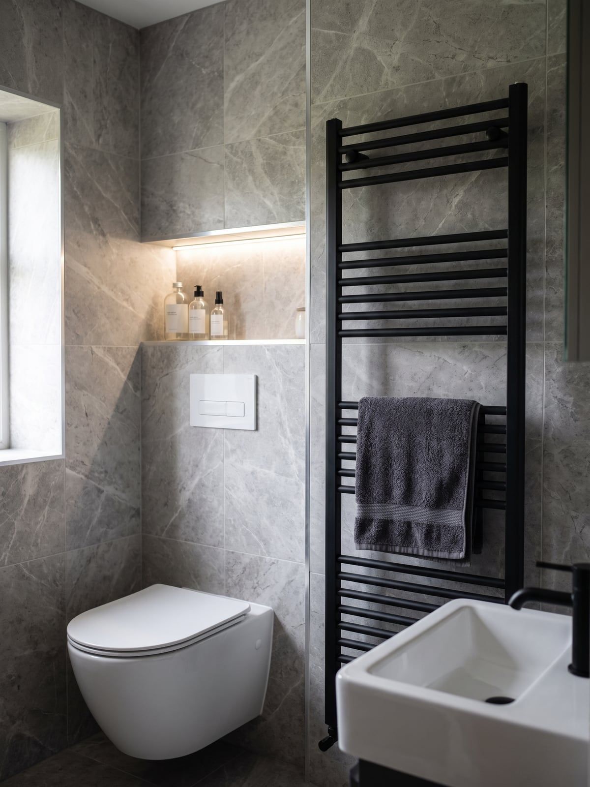 Wall-hung WC against grey marble-effect tiles, with a lit shelf recess above it and a matt black towel radiator beside it