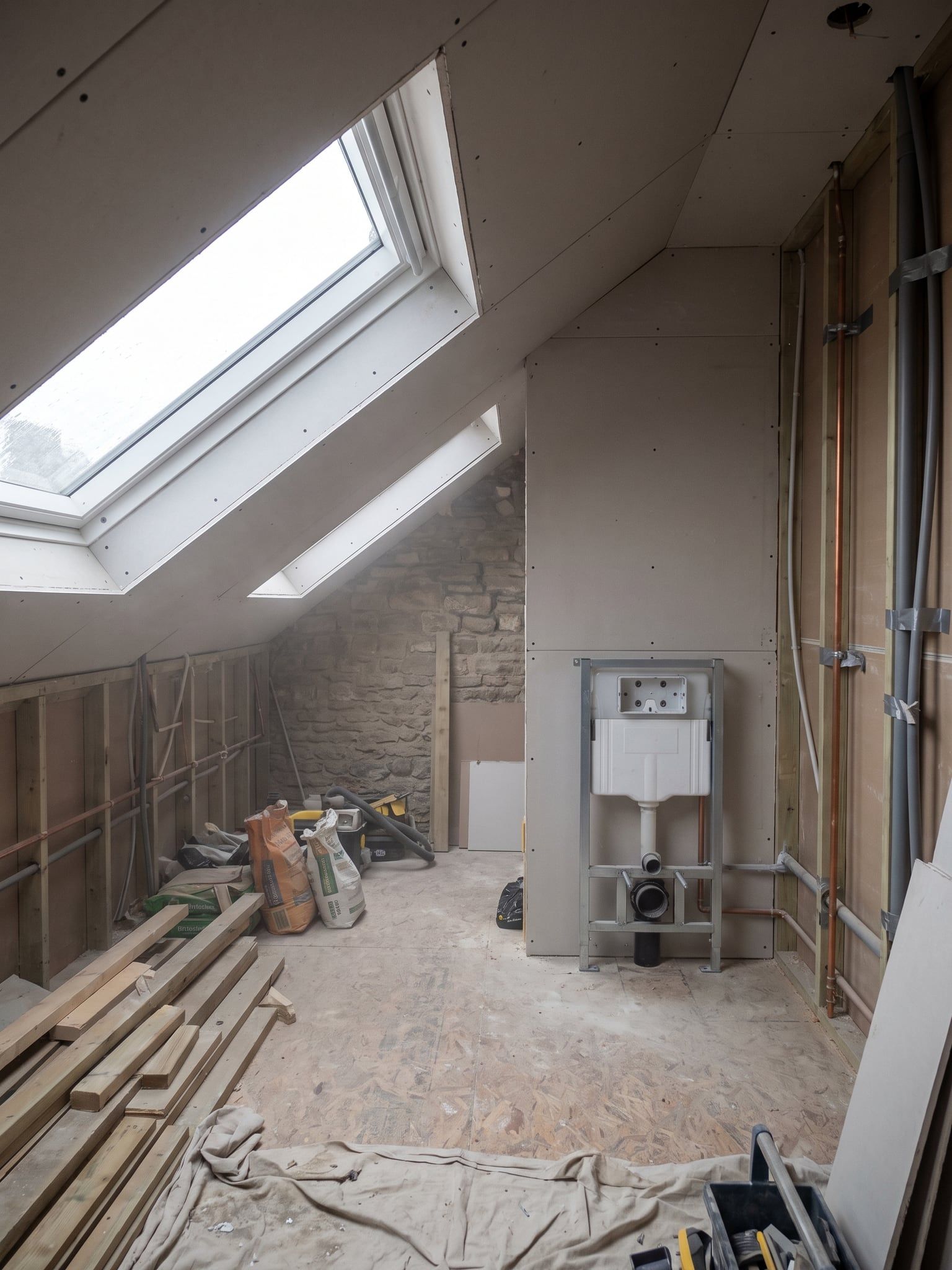 Bare loft room during the renovation, with loose pipework, a WC mounting frame and building materials on the floor
