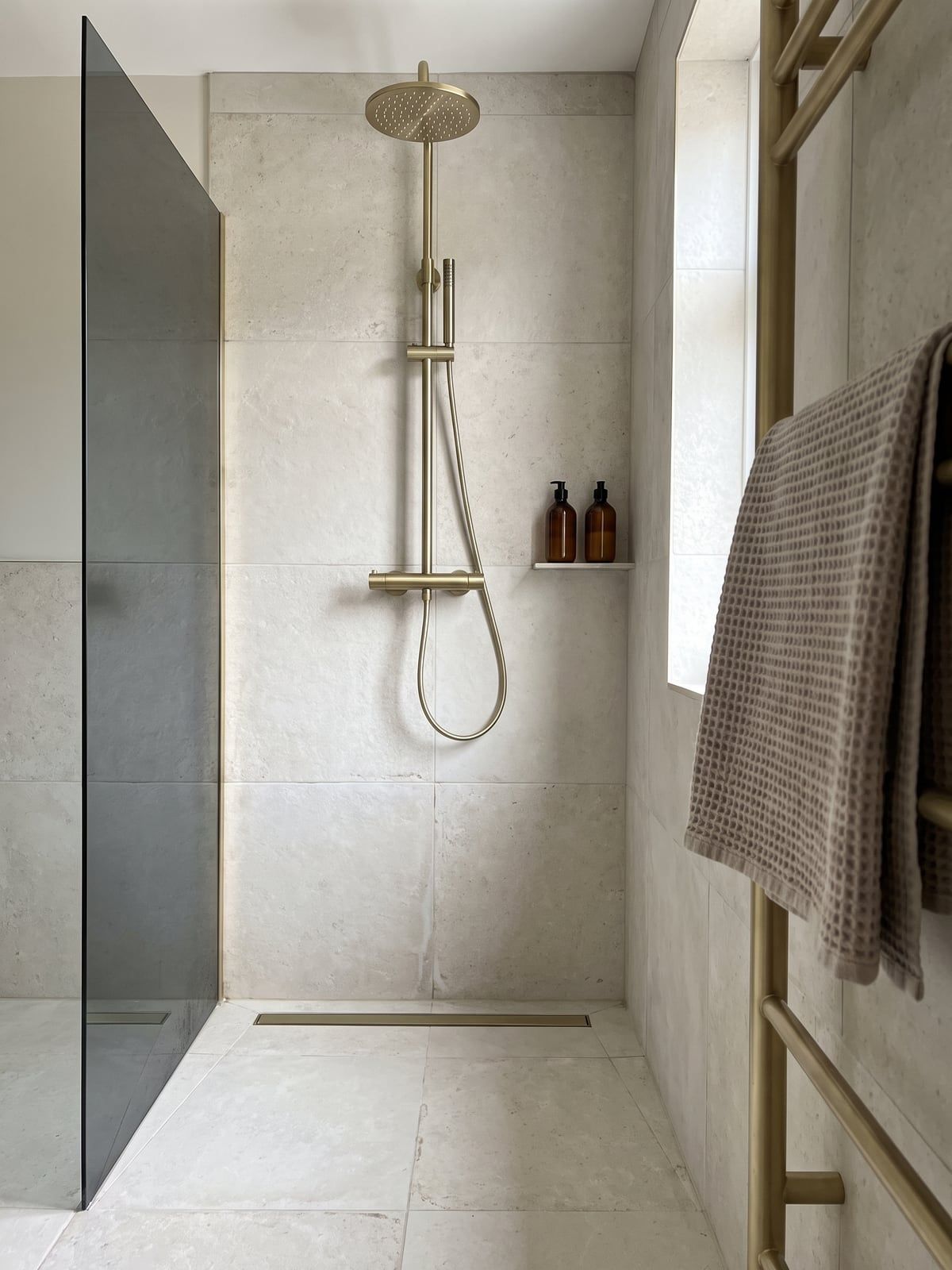 Walk-in shower with a brushed brass rain shower on a tall riser, a smoked glass screen and a long linear floor drain